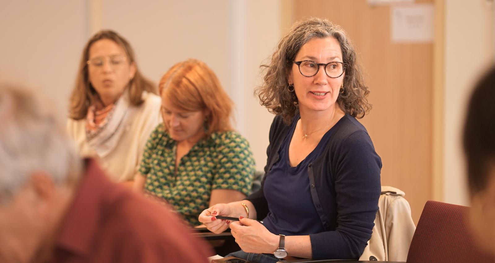 A woman seated in a chair during a conference, hands placed on her lap and looking to her left as in conversation.
