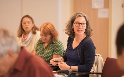 A woman seated in a chair during a conference, hands placed on her lap and looking to her left as in conversation.