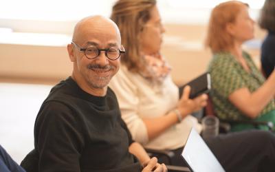 A man with glasses is seated in a chair, focused on his laptop during a conference and smiles.