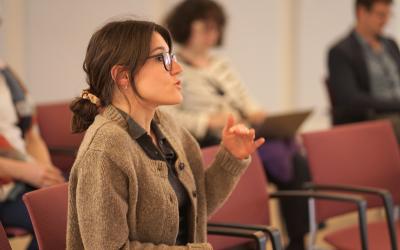 A woman in glasses is sitting in a chair, in the middle of a conversation during a conference.