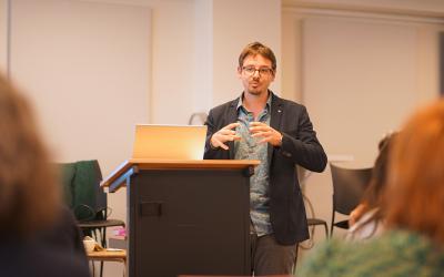 A man presenting to an audience, gesturing as he explains his topic to the engaged group of listeners.