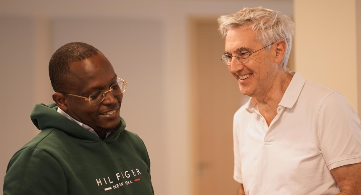 Two men wearing a white shirt and a green hoodie talking to each other in a casual setting.