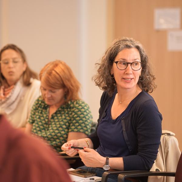 A woman seated in a chair during a conference, hands placed on her lap and looking to her left as in conversation.