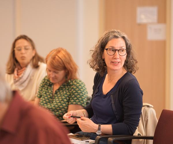 A woman seated in a chair during a conference, hands placed on her lap and looking to her left as in conversation.